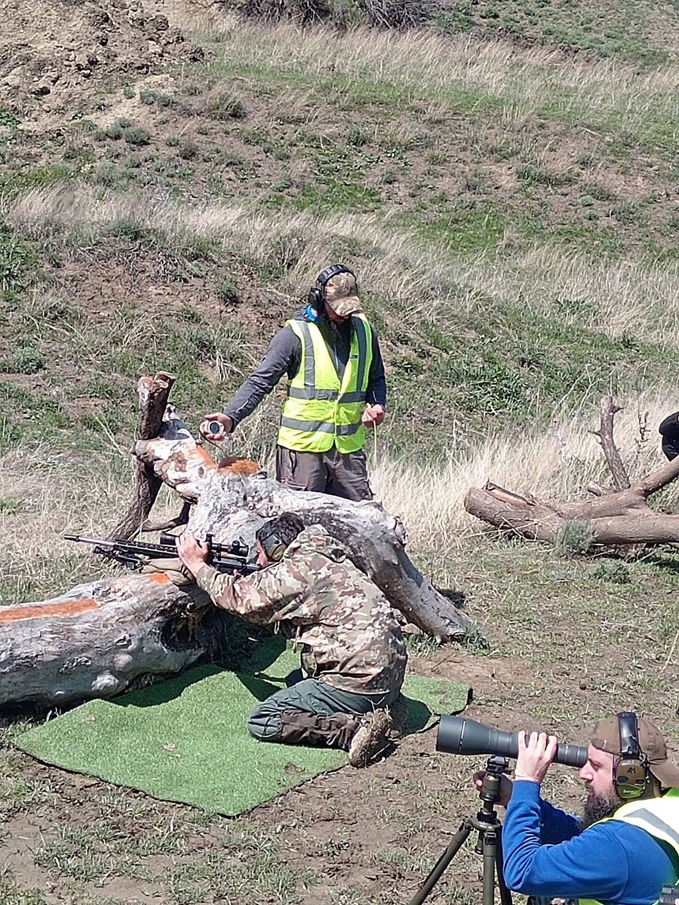 A shooter firing off a log prop while the range officer and a spotter watch