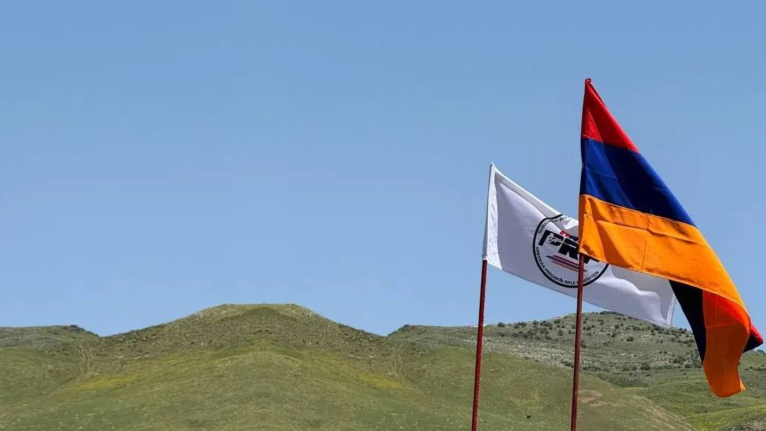 The Armenian flag and the ARMPRF flag over the range, green hills behind them